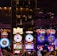 A lively casino scene featuring a row of bright, colorful slot machines with themes like Monopoly, Michael Jackson, and Lord of the Rings. The gaming machines display a variety of flashing lights and digital screens, creating an energetic and exciting atmosphere. Reflections of the lights are visible in glossy surfaces above the machines.